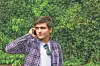 A man in a plaid shirt talks on a phone in front of green foliage.