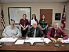 Five officials seated behind a conference table with papers, nameplates, maps, and flags in a government office.