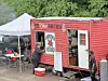 A red food truck parked next to a white tent at an outdoor event with people nearby.