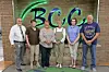 Six people standing side by side in front of a brick wall with a large green and black sign.