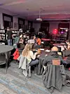 People seated at tables in a lounge with a bar, dark furniture, and red lighting in the background.