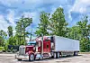 Red semi-truck with a silver trailer parked in a lot beside trees.