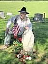 Man sitting beside a grave marker with flowers and an American flag in a cemetery.