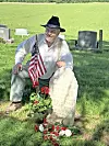Man sitting beside a grave marker with flowers and an American flag in a cemetery.
