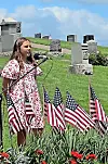 Girl speaking into a microphone at a cemetery with rows of American flags and gravestones behind her.