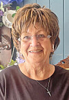 Smiling older woman wearing glasses and a cross necklace in front of a floral arrangement.