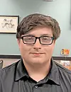 Person wearing glasses and a dark shirt indoors with framed wall art behind them.