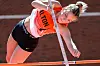 Athlete in orange and black clears the pole vault bar during a track and field event.