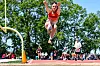 Athlete in orange jumps on a track and field runway with spectators and trees in the background.