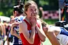 Female track athlete in a red uniform covering her mouth after a race, with other runners behind her.