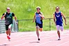 Three runners sprint on an outdoor track with a grassy field in the background.