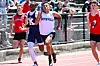 Three runners sprint on a track while spectators watch from the stands.