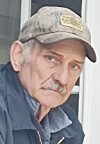 Man in a camouflage cap and dark jacket standing near a door, looking toward the camera.
