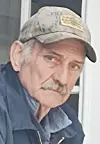 Man in a camouflage cap and dark jacket standing near a door, looking toward the camera.