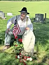 Man seated beside a grave marker with an American flag and red flowers in a cemetery.