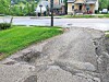 Cracked asphalt path with potholes beside a grassy area and houses across the street.
