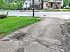 Cracked asphalt path with potholes beside a grassy area and houses across the street.