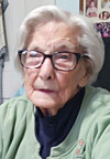 Elderly woman with glasses wearing a green sweater seated indoors.