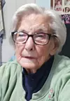 Elderly woman with glasses wearing a green sweater seated indoors.