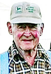 Portrait of an elderly man wearing a white cap, plaid shirt, and blue vest against a light background.