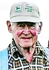 Portrait of an elderly man wearing a white cap, plaid shirt, and blue vest against a light background.