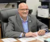 A smiling man sits at an office desk with paperwork and a pen, with office equipment behind him.