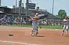 Two softball players hug on the infield during a game, with another player standing to the right and spectators behind them.