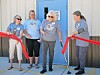 Four women stand by a red ribbon outside a building entrance during a ribbon-cutting ceremony.