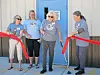 Four women stand by a red ribbon outside a building entrance during a ribbon-cutting ceremony.