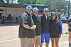 Four people stand together on a baseball field holding a yellow ball, with a dugout and spectators behind them.