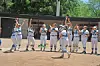 Youth baseball players in uniform stand on a dirt field with their arms raised.