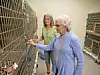 Two women stand in a shelter aisle beside metal cages containing animals.