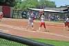 Youth softball players stand on the infield during a game with spectators behind the fence.