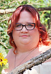 Woman with red hair and glasses standing outdoors near branches and flowers.