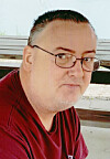 A man in glasses and a red shirt sits outdoors near a bench.