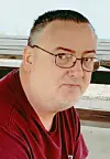 A man in glasses and a red shirt sits outdoors near a bench.