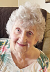 Elderly woman with curly white hair seated on a couch and facing the camera.