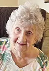 Elderly woman with curly white hair seated on a couch and facing the camera.