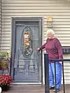 Older woman standing on a porch next to a decorated front door.