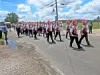 A marching band marches along a road beside a puddle under a partly cloudy sky.
