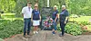 Four people standing beside a memorial plaque and flowers in a cemetery.