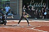 A softball batter readies a swing at home plate while spectators stand behind the fence.