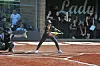 A softball batter readies a swing at home plate while spectators stand behind the fence.