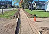 A gravel trench beside a residential street with an orange traffic cone and houses in the background.