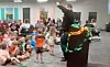 A speaker at a podium addresses a seated crowd in a room with children and adults.