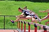 Track athletes jumping over hurdles during a race on a red running track.
