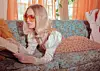 Woman in sunglasses reading a book while lying on a floral sofa near orange curtains.