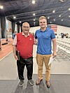 Two men standing on an indoor track beside rows of chairs, with an American flag hanging in the background.