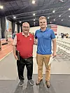Two men standing on an indoor track beside rows of chairs, with an American flag hanging in the background.