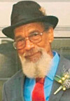 Close-up portrait of an elderly man wearing a black hat, glasses, and a suit with a red tie.